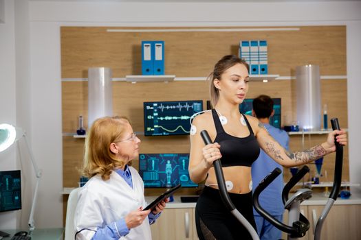 Female athlete making physical effort on stepper and holding electrodes on her and doctor holding tablet in hand. Tests to run