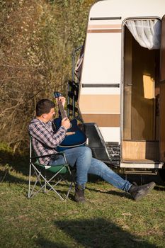 Young man singing music on his guitar in the mountains in front of his retro camper van.