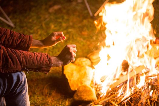 Young man warming up his hands above the bonfire. Campsite in the mountains.