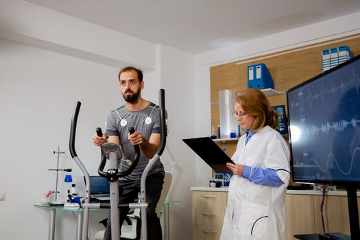 Male athlete who makes a physical effort on the stepper and has electrodes attached to it and the doctor makes notes on the clipboard.