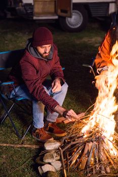 Bearded tourist in the mountains making camp fire in a cold night of autumn. Retro camper van.