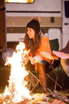 Beautiful young girl laughing and warming around camp fire in a camp site with retro camper van in the background.