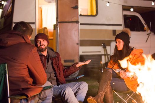 Group of friend enjoying clean air in the mountains and sharing a story around camp fire. Retro camper van.