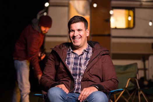 Portrait of young man smiling with retro camper van in a camp site in the mountains. Light bulbs.