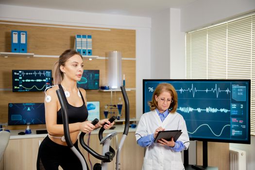 Female athlete testing the stepper with electrodes on her and the doctor making notes in the tablet. Sports laboratory