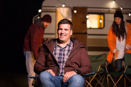 Tourist in a camp site smiling while his friends are making camp fire. Retro camper van.