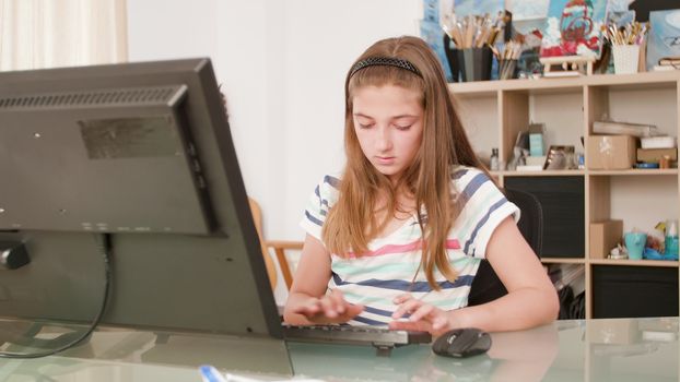 Young girl types a text at a keyboard and uses the mouse. Girl doing her homework in her room.