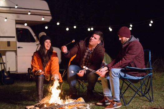 Group of friends laughing while roasting marshmallows on camp fire. Retro camper van.