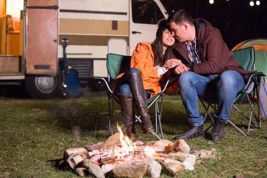 Couple showing affection to each other near camp fire in a cold night of autumn. Retro camper van the background.