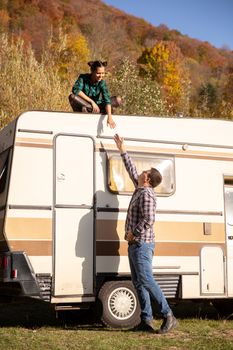 Man trying to lend a hand to his girlfriend sitting on the roof of a retro camper. Autumn colors