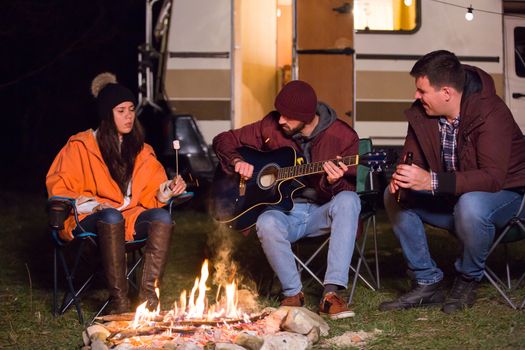 Young friends roasting marshmallows over camp fire and playing songs in guitar. Retro camper van.