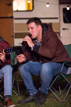 Young man enjoying a bottle of beer in a cold night of autumn in a campsite. Retro camp fire.