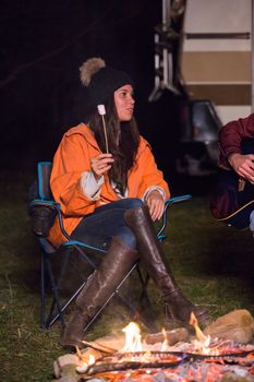 Cheerful young woman holding a roasted marshmallow near camp fire with retro camper van in the background.