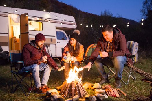 Close friends drinking beer together in the mountains and warming their hands around camp fire. Retro camper van with light bulbs.