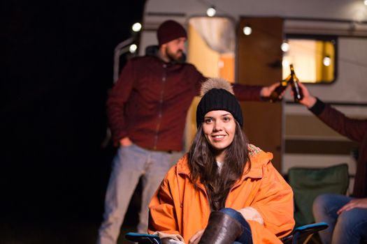 Portrait of young girl in front of retro camper van with friends clinking beer in the background.