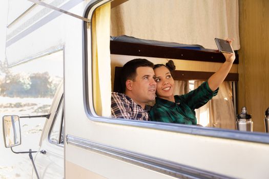 Beautiful young woman with a big smile inside retro camper van taking a selfie with her boyfriend while enjoying their vacation.