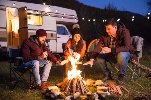 Group of friends enjoying the fresh air of the mountains around camp fire. Retro camper van with light bulbs.