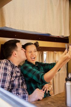 Beautiful young girlfriend making a funny grimace while taking a selfie. Boyfriend kissing his girlfriend cheek while taking a selfie. inside retro camper van.