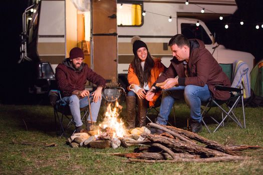 Group of friend relaxing and laughing together around camp fire in the mountains. Retro camper van.