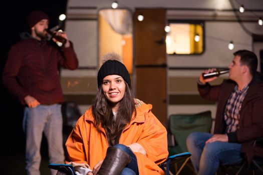 Portrait of young girl in front of camp fire with friends drinking beer in the background. Retro camper van with light bulbs.