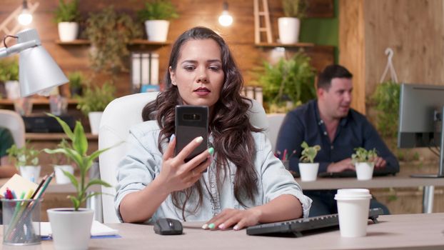 Woman has a video conference on her phone in a cozy office. Medium shot. Her colleagues are working in the background