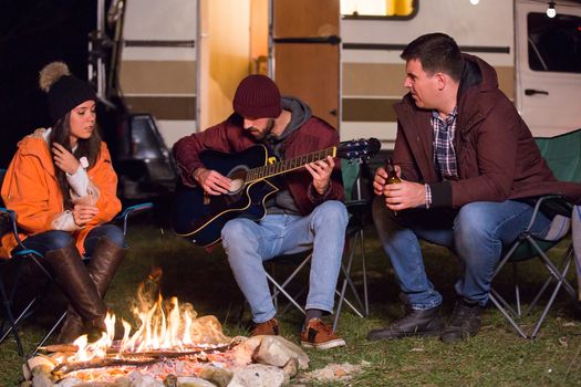 Friends relaxing and playing on guitar around camp fire with their retro camper van in the background. Light bulbs.