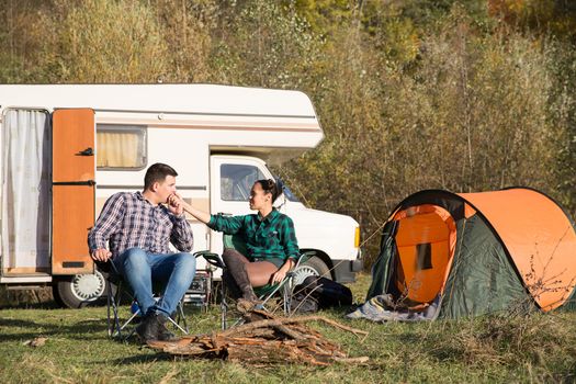 Boyfriend kissing the hand of his girlfriend on camping site with retro camper van in the background.