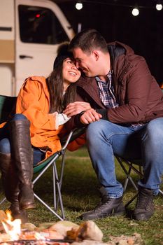 Couple smiling and enjoying the warmth of camp fire in a cold night of autumn in the mountains with retro camper van in the background.