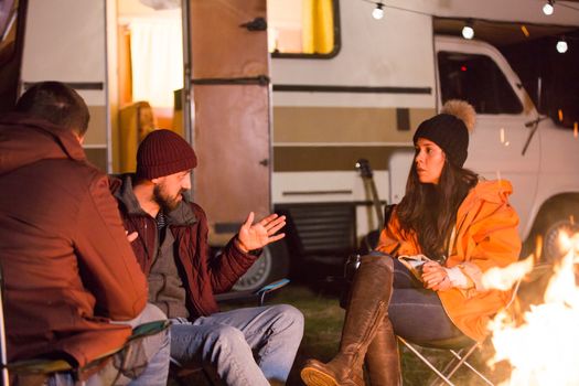 Bearded man telling a story to his friends around camp fire in a cold night of autumn in the mountains. Retro camper van.