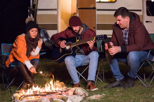 Caucasian girl roasting marshmallow over camp fire and her friend is playing on guitar with their retro camper van in the background.