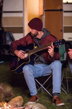 Passionate young man playing on guitar for his friends around camp fire with their retro camper van in the background.