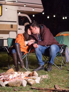 Couple sitting on camping chairs in a cold night of autumn near warm camp fire. Retro camper van with light bulbs.