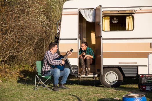 Carefree man playing on guitar for his girlfriend in front of their retro camper van. Couple relaxing in the mountains.