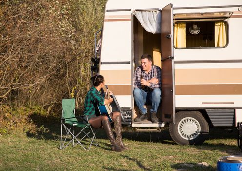 Woman playing on old guitar for his man in front of their retro camper van in the mountains.