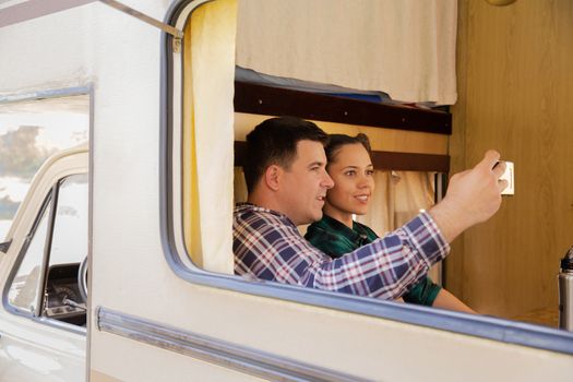 Beautiful young couple inside their retro camper van taking a selfie in a campsite.