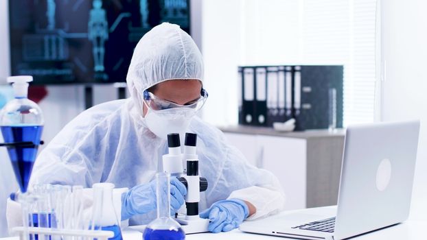 Chemist in coverall equipment typing formula on laptop. Scientist doing lab analysis using microscope.