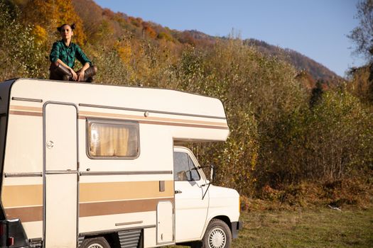 Caucasian girl sitting on the roof of a camper in a beautiful autumn landscape. Recreation travel