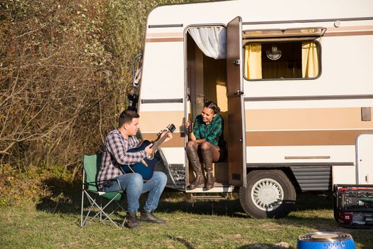 Man trying to play a song on old guitar for his woman in front of their retro camper van. Couple in the mountains.