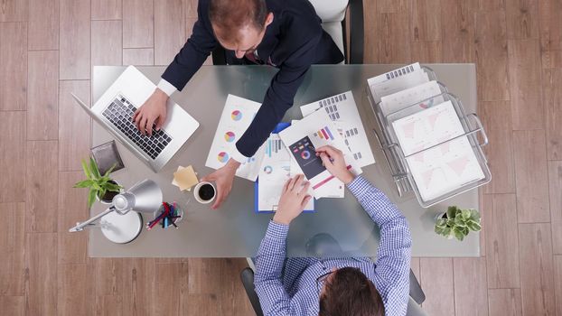 Top view of businessman in suit drinking a cup of coffee while his consultant explaning charts sitting in the office.