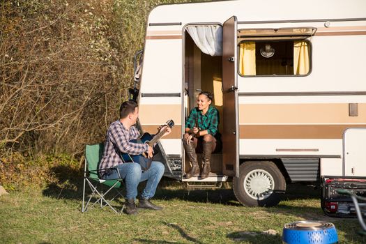 Cheerful woman relaxing and listening how boyfriend is playing on their old guitar in front of their retro camper van.
