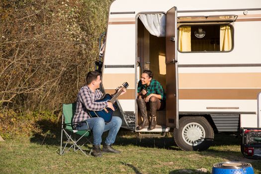 Woman smiling and looking at her hipster boyfriend playing on old guitar in front of their retro camper van.