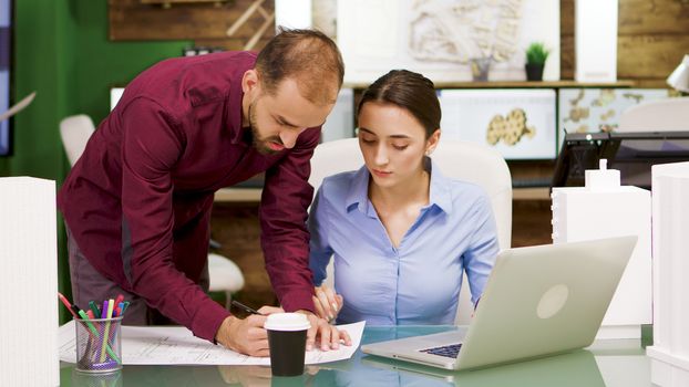 Female chief architect taking a sip of coffee and helping her colleague with a project on blue prints. Architect working on laptop.