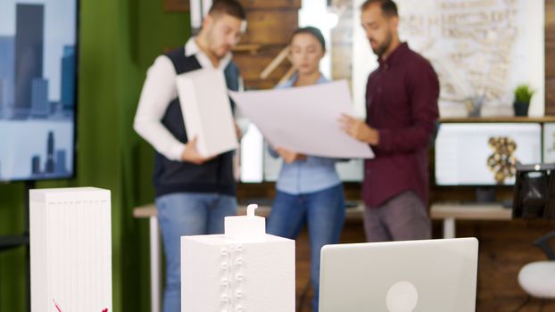 Slide view of building models with architects working in the background. Architect holding blue print.