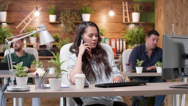 Woman talks on the phone in a busy cozy office. Her colleagues are working in the background