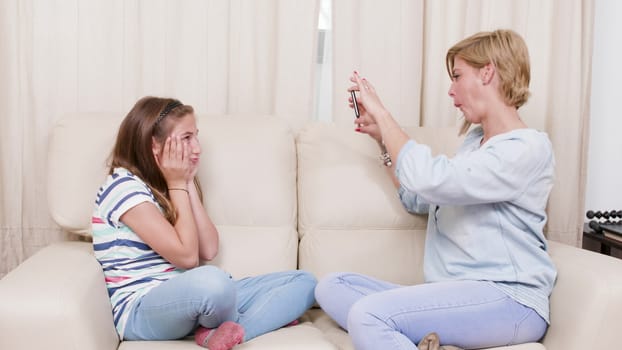 Mother taking pictures of her daughter while relaxing on a sofa. Mother and her daughter making photos on a smartphone.