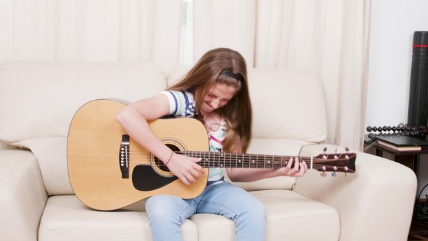 Teenage girl plays on an acoustic guitar sitting on a sofa. Teen playing on a musical instrument.