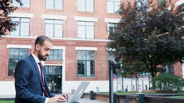 Businessman in formal suit outdoor typing on the laptop sitting in a business district. Panning down shot