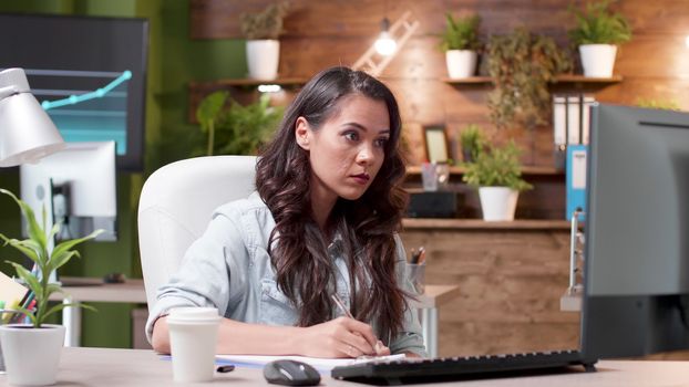 Concentrated woman reads an email and takes notes on the clipboard at her desk. Modern cozy office. Her colleagues are working in the background