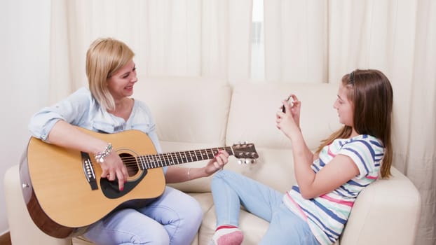 Teenage girl taking pictures of her mother playing on an acoustic guitar. Mother and daughter having fun in the living room.