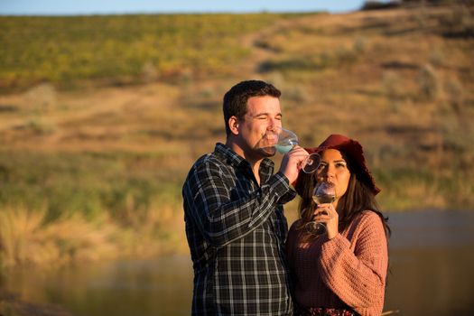 Wine grower couple enjoying a glass of wine in a vineyard with lake behind them at the sunset.